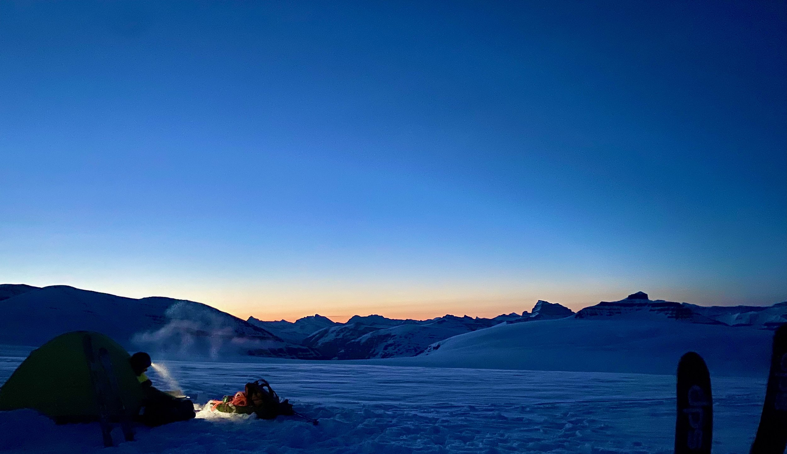 Alpenglow over snow-covered peaks at dusk, with a tent and skis in the foreground